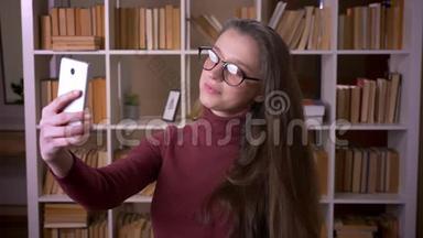 <strong>戴</strong>眼镜的年轻白种人女学生在大学图书馆室内通过<strong>手机自拍</strong>的特写照片
